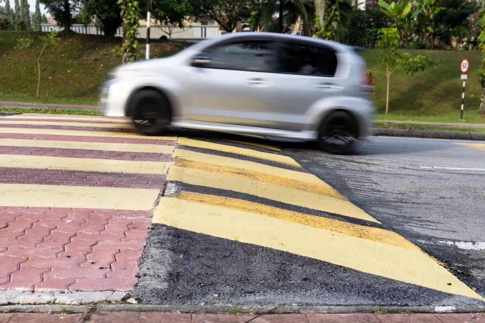 Small silver car driving over wide soeed cushion on asphalt road.